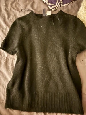 Svart stickad topp med kort ärm - En svart stickad topp med korta ärmar och rund hals. Toppen har en enkel, klassisk design med ribbad kant nedtill. Perfekt att styla med jeans eller kjol för en clean look.