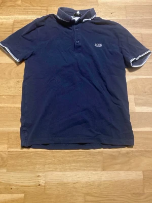 Marinblå pikétröja från BOSS - Snygg marinblå pikétröja från BOSS med vit logga på bröstet och kontrasterande vita detaljer på krage och ärmslut. Klassisk pikéknäppning framtill och korta ärmar. Perfekt till en avslappnad men stilren look.