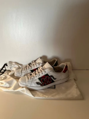 Vita sneakers med ormtryck - Säljer ett par vita sneakers med coolt ormtryck på sidan och detaljer i rött och grönt. Skorna har klassisk låg profil och vita snören. Materialet är slätt läder och sulan är i gummi. Perfekta för dig som vill sticka ut med din stil.