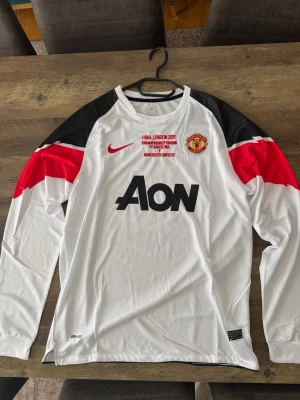 Manchester United långärmad finaltröja - Vit långärmad Manchester United-tröja från Nike med svarta och röda detaljer. Tryck på bröstet för Champions League-finalen 2011 mot Barcelona. Broderad klubbmärke och AON-logga på framsidan. Tillverkad i lätt och ventilerande material.