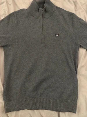 Grå stickad halvzip tröja från Lexington - Mörkgrå stickad tröja med halvzip och hög krage. Liten broderad logga på bröstet. Tröjan har ribbade muddar vid ärmslut och nederkant. Perfekt för lager-på-lager och enkel att matcha med jeans eller chinos. Bara att höra av er om prisförslag