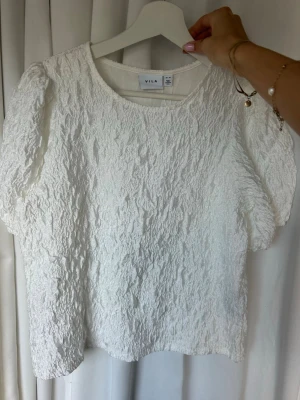Vit strukturerad topp från Vila - Säljer en vit topp från Vila i storlek XS. Toppen har en unik strukturerad yta och korta ärmar. Den har rund halsringning och är tillverkad i ett lätt material som ger en cool look. Perfekt att styla med jeans eller kjol.