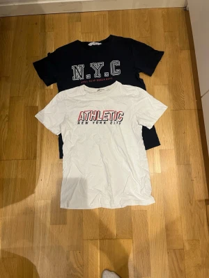2-pack t-shirts från H&M - Säljer två t-shirts, en svart med stort 'N.Y.C' tryck och en vit med 'ATHLETIC New York City' tryck. Båda har klassisk passform, rund hals och korta ärmar. Perfekta för en avslappnad streetwear-look.