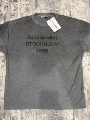 Grå Acne Studios t-shirt - Snygg grå t-shirt från Acne Studios med tryck 'STOCKHOLM 1996' på bröstet. Klassisk rund hals och korta ärmar. Perfekt basplagg med stilren design och mjukt material.
