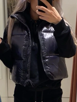 Svart glansig dunväst med dragkedja - Snygg svart dunväst med glansig finish och hög krage. Västen har en rak passform, dragkedja framtill och justerbar dragsko nedtill. Perfekt att slänga över en hoodie för en trendig lager-på-lager-look.