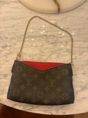 Monogram handväska med kedjerem - Snygg handväska brunt monogrammönster med guldfärgad kedjerem och röd detalj upptill. Väskan har en elegant och kompakt form, perfekt för att bära det viktigaste. Materialet är tåligt canvas med läderdetaljer. Inte äkta. Vid intresse skriv för mer info om väskan