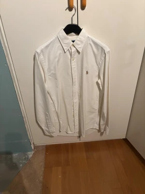 Vit klassisk skjorta med broderad logga - Klassisk vit skjorta i bomull med lång ärm och knappar framtill. Skjortan har en broderad logga på bröstet och en stilren krage. Perfekt till både jeans och kostymbyxor för en clean look.
