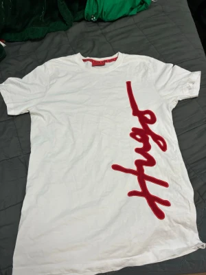Vit t-shirt från HUGO med röd text - Snygg vit t-shirt från HUGO med ett stort rött broderat tryck längs framsidan. Klassisk rund halsringning och korta ärmar. Tillverkad i mjuk bomull för skön känsla och stilren look storlek S men passar bra på m