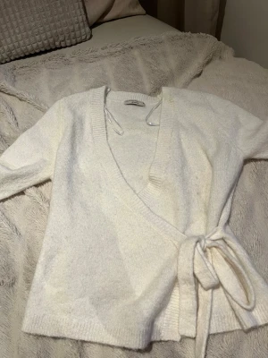 Vit omlottkofta från Gina Tricot - Säljer en mjuk och stilren vit stickad omlottkofta från Gina Tricot. Koftan har knytband i midjan, ribbade kanter och lång ärm. Perfekt att slänga på sig för en avslappnad men snygg look.