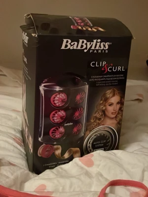 BaByliss Clip & Curl värmespolar - BaByliss Clip & Curl värmespolar med auto-clip system för enkel styling. Innehåller 12 värmespolar i två olika storlekar, perfekta för att skapa mjuka lockar och volym. Spolarna är svarta och rosa och kommer i en praktisk förvaringsbox.