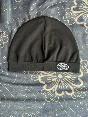 Svart tunn mössa med broderad logga - Säljer en svart, tunn och stretchig mössa med en broderad vit logga framtill. Mössan har enkel design och ribbad kant nedtill. Perfekt för dig som vill ha en stilren accessoar till hösten eller vintern.