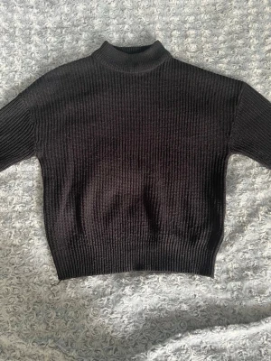 Svart ribbstickad tröja med hög krage - En svart, ribbstickad tröja med hög krage och långa ärmar. Tröjan har en croppad passform och breda muddar vid ärmslut och nederkant. Perfekt för lager på lager och enkel att matcha med olika stilar.