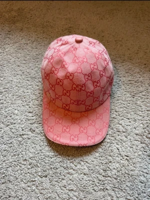 Gucci keps  - Säljer en snygg rosa keps från Gucci med det klassiska GG-monogrammet över hela kepsen. Kepsen har böjd skärm och är tillverkad i ett slitstarkt tygmaterial. Perfekt för dig som vill ha en trendig och exklusiv accessoar till din outfit.