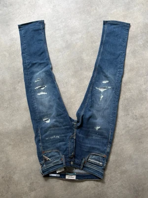 Replay jeans med slitningar - W27 men sitter som 28/29. Säljer ett par blå jeans med raka ben och slitna detaljer framtill. Jeansen har klassisk femficksdesign, normal midja och dragkedjegylf. Materialet är denim i bomull med en snygg tvättad look och slitningar för en trendig vibe.