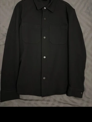 Svart overshirt  - Stilren svart overshirt från Zara med klassisk krage, två bröstfickor och knäppning framtill. Materialet är mjukt och känns bekvämt, perfekt att slänga på sig över en t-shirt, tröjor och även hoodies. Passar dig som gillar minimalistisk och clean look. 