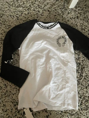 Chrome Hearts svartvit långärmad tröja - Säljer en svartvit långärmad tröja från Chrome Hearts med kontrastärmar och stora logotryck på både bröstet och ryggen. Tröjan har även tryck längs ärmarna och logga i halsen. Perfekt för dig som gillar streetwear och vill sticka ut.
