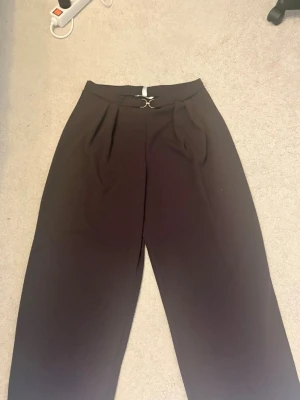 Bruna vida kostymbyxor med metallspänne - Snygga bruna kostymbyxor med hög midja och vida ben. Byxorna har ett dekorativt metallspänne framtill och veckade detaljer vid midjan. Perfekta för en stilren och trendig look. Materialet ser ut att vara mjukt och följsamt.