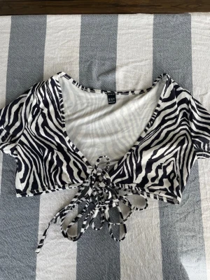 Zebramönstrad crop top från SHEIN - Snygg zebramönstrad crop top från SHEIN med korta ärmar och knytning framtill. Toppen har djup v-ringning och är i ett mjukt, stretchigt material som sitter bekvämt. Perfekt för att skapa en trendig och cool look.