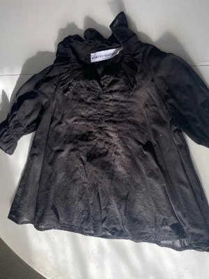 Svart blus med volangkrage - Snygg svart blus från Howtokissafrog med volangkrage och knappar framtill. Blusen har trekvartsärm med resår vid ärmslut och är tillverkad i ett lätt tyg som ger ett avslappnat intryck. Perfekt för dig som gillar en trendig och unik look.