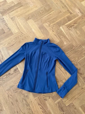 Marinblå träningsjacka med dragkedja - Snygg marinblå träningsjacka med hel dragkedja framtill och två fickor med dragkedja. Figurnära passform och hög krage. Perfekt för löpning eller gymmet. Materialet känns stretchigt och bekvämt.