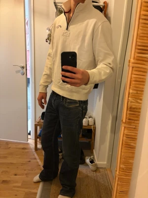 Vit halvzip tröja - Säljer en stilren vit halvzip tröja med broderad detalj på bröstet. Tröjan har hög krage, muddar vid ärmslut och nederkant samt en avslappnad passform. Perfekt för dig som gillar en clean och enkel look.