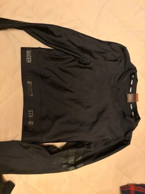 Svart träningsset från Stronger - Snyggt svart träningsset från Stronger med långärmad topp och matchande tights. Setet har diskreta loggor och detaljer i svart, samt stretchigt material som passar perfekt för gym eller löpning. Både tröjan och tightsen har en tight passform.