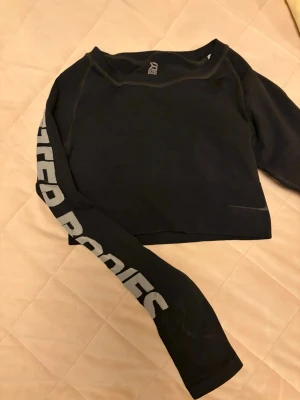 Svart croppad långärmad topp - Svart croppad långärmad topp med tryck i vitt längs ena ärmen. Perfekt till gymmet eller som en cool vardagslook. Rund halsringning och mjukt material som känns skönt mot huden.