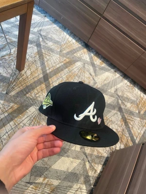 Svart Atlanta Braves keps - Säljer en svart snapback-keps med Atlanta Braves-logga i vitt framtill och broderad 1995 World Series-detalj på sidan. Kepsen har platt skärm och små broderade hjärtan vid loggan. Klassisk streetwear-stil, perfekt för dig som gillar sportiga accessoarer.