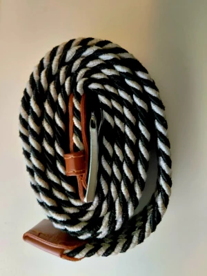 Onewear bälte - Säljer ett svartvitt randigt flätat bälte med bruna läderdetaljer och silverfärgat spänne. Bältet har en snygg twistad design som ger en cool look till outfiten. Perfekt för dig som vill ha något som sticker ut lite extra.
