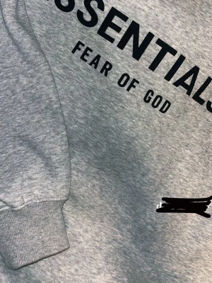Grå Essentials Fear of God hoodie - PRIS KAN DISKUTERAS Snygg grå hoodie från Essentials Fear of God med svart logga tryckt på bröstet och ryggen. Klassisk känguruficka framtill, ribbade muddar och huva. Tillverkad i mjukt bomullsmaterial som är perfekt för en avslappnad streetwear-look.