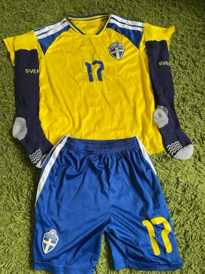 Sverige fotbollsset nr 17 - Säljer ett komplett Sverige fotbollsset med gul tröja och blå shorts, båda med nummer 17 och landslagets emblem. Tröjan har blå och vita detaljer samt namnet Gyökeres på ryggen. Ingår även ett par blå fotbollsstrumpor med texten 'Sverige'. Perfekt för match eller träning!