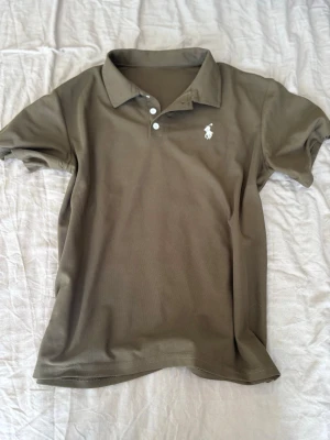 Olivgrön pikétröja från Ralph Lauren - Snygg olivgrön pikétröja från Ralph Lauren med klassisk krage, knappar framtill och den ikoniska vita broderade loggan på bröstet. Perfekt till jeans eller shorts för en clean och stilren look.