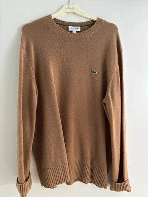 Brun stickad tröja från Lacoste - Säljer en klassisk brun stickad tröja från Lacoste med rund halsringning och ribbade muddar. Tröjan har den ikoniska gröna krokodilloggan på bröstet och är i en mjuk, skön kvalitet. Perfekt för dig som gillar stilrena och tidlösa plagg.