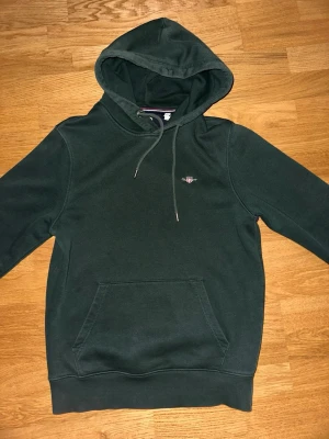 Mörkgrön hoodie från Gant - Mörkgrön hoodie från Gant med broderad logga på bröstet. Klassisk modell med huva, dragsnören och stor magficka framtill. Mjuk bomullskänsla och ribbade muddar vid ärmslut och nederkant. Perfekt för en avslappnad stil.