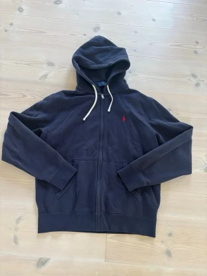 Mörkblå hoodie med dragkedja Polo Ralph Lauren - Mörkblå hoodie från Polo Ralph Lauren med dragkedja framtill och vit dragsko i huvan. Klassisk röd logga broderad på bröstet. Två fickor framtill och ribbade muddar vid ärmar och nederkant. Perfekt för en avslappnad och stilren look.              En defekt under ena armen, lagat hål inte synligt