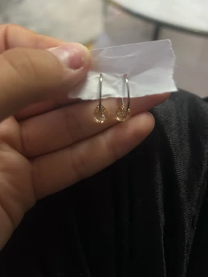 Guldiga örhängen med glittrande sten - Snygga örhängen i guldton med rund glittrande sten som hänger i tunn båge. Perfekta för dig som gillar stilrena och eleganta accessoarer. Materialet ser ut att vara metall med en klar sten som detalj. Nytt skick (aldrig använt)