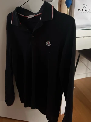 Svart långärmad Moncler - Svart långärmad polotopp från Moncler med klassisk krage och knäppning framtill. Perfekt till jeans eller kjol för en clean look. Skick 8/10 ingen defekt med hål eller nån sån skit. Ny pris 3600kr. MVH ELIAS!