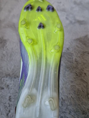 Adidas F50 lila och neongröna fotbollsskor - Säljer ett par Adidas F50 fotbollsskor i lila och neongrönt. Skorna har FG-dobbar för spel på naturgräs och en strumpliknande ovandel för skön passform. Lätta material och modern design med tydliga F50-detaljer och Adidas-logga.
