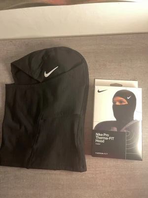 Nike Pro Therma-FIT svart balaclava - Nike Pro Therma-FIT balaclava i svart funktionsmaterial. Huva med vit Nike-logga framtill, designad för att täcka huvud, hals och ansikte. Perfekt för träning utomhus eller kalla dagar. Snygg och sportig look.