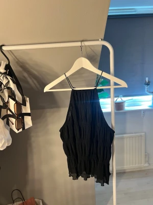 Svart halterneck topp med tunna band - Trendig svart halterneck topp med tunna axelband och vågig, lätt skrynklig struktur. Toppen har en lös passform och faller snyggt med volangkant nedtill. Perfekt för dig som gillar en stilren men ändå unik look.