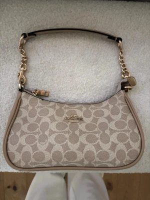 Beige axelremsväska från Coach - Snygg beige handväska från Coach med klassiskt monogrammönster och guldfärgade detaljer. Väskan har en kort axelrem med kedja och läder, samt dragkedja upptill. Perfekt för dig som gillar stilrena accessoarer med lyxig känsla.