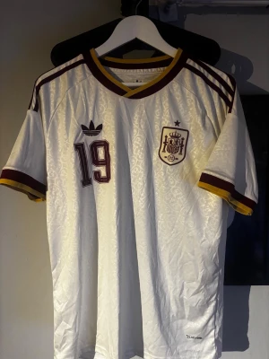 Vit Spanien fotbollströja nr 19 - Säljer en vit Spanien fotbollströja med vinröda och gula detaljer. Tröjan har nummer 19 och namnet Lamine Yamal på ryggen. Klassisk V-ringning med randiga muddar och broderad landslagssköld på bröstet. Tillverkad i lätt och ventilerande material.