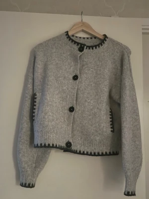 Grå stickad kofta med svarta detaljer - Mysig grå stickad kofta med stora svarta knappar och svarta detaljer längs kanterna. Rak passform, rund halsringning och långa ärmar. Perfekt att slänga över en t-shirt eller topp för en avslappnad look.