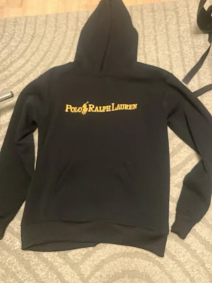 Svart hoodie från Polo Ralph Lauren - Snygg svart hoodie från Polo Ralph Lauren med gul logga och text på bröstet. Klassisk huva och magficka framtill. Perfekt för dig som vill ha en stilren och bekväm hoodie med ikonisk design.