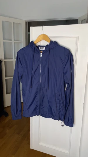 Blå vindjacka med huva från RISKY - Snygg blå vindjacka från RISKY med dragkedja framtill, justerbar nederkant och två fickor. Jackan har huva med tryckt logga på baksidan och tunnt, lätt material som passar perfekt till blåsiga dagar.