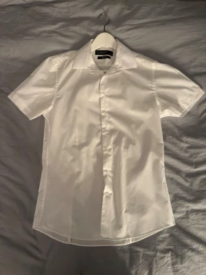 Vit kortärmad skjorta - Klassisk vit skjorta med korta ärmar och knappar framtill. Skjortan har en enkel, stilren design med krage och är tillverkad i ett lätt bomullstyg som passar perfekt till sommaren.