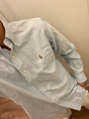 Ljusblå skjorta från Polo Ralph Lauren (Slim Fit) - Klassisk ljusblå skjorta från Ralph Lauren med broderad logga på bröstet. Skjortan har knappar framtill, button-down krage och långa ärmar. Tillverkad i mjuk bomull för en bekväm känsla och tidlös stil. Storlek M Slim fit. Snabb Leverans!