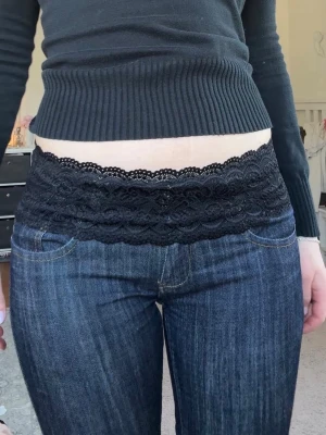 Svart spetsig midjeband  - Svart midjeband i stretchig spets med vågig kant och blommigt mönster. Perfekt att ha  på jeans för en snygg lager-på-lager-look. Materialet är mjukt och elastiskt för bekväm passform.