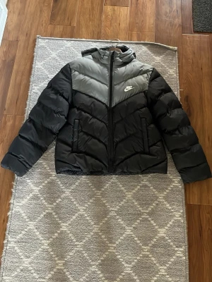 Svart och grå pufferjacka från Nike - Snygg pufferjacka från Nike i svart och grått med huva och dragkedja framtill. Jackan har ett quiltat mönster och logga på bröstet. Perfekt för kalla dagar när du vill hålla stilen och värmen.