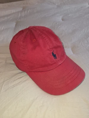 Röd keps från Polo Ralph Lauren - Snygg röd keps från Polo Ralph Lauren med klassisk böjd skärm och blå broderad logga framtill. Tillverkad i 100% bomull för en bekväm och luftig känsla. Justerbar baktill för perfekt passform. Är liten i storleken för att de är en barn keps 